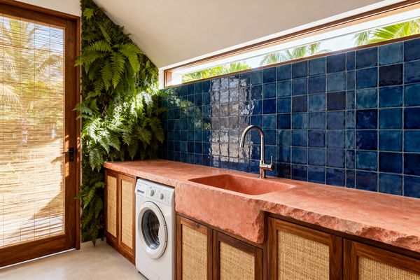 Bright laundry room with a coral-pink stone countertop and integrated sink set against glossy indigo square tiles. Warm wood cabinets with woven rattan fronts frame a white front-loading washer, while a vertical garden of ferns and a wood door with a bamboo shade filter sunlight; a slim clerestory window above the backsplash reveals palm trees outside.