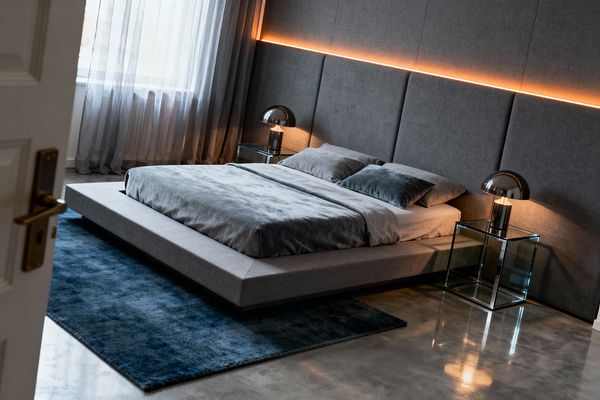 A contemporary bedroom features a low gray upholstered platform bed set against large gray fabric headboard panels. A thin horizontal strip of warm LED light runs across the headboard. Matching glass cube nightstands hold chrome dome table lamps. A deep navy rug softens a polished concrete floor, while floor-to-ceiling sheer gray curtains filter daylight from a large window, creating a minimal, symmetrical, and softly lit space.