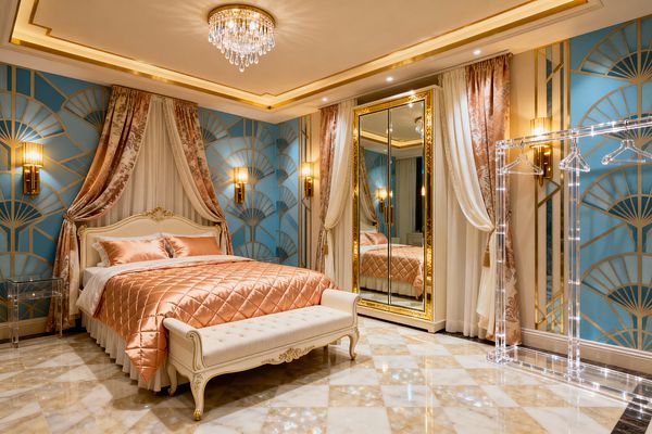 A luxurious kids’ bedroom with an ivory carved bed dressed in blush satin quilt and pillows, framed by layered ivory sheers and rose damask drapery. Powder-blue walls feature gold Art Deco fan patterns with brass sconces. A tufted bench rests on white-and-beige marble checkerboard flooring. A mirrored wardrobe with gilded frame reflects a crystal chandelier and warm cove lighting, while a clear acrylic garment rack with hangers stands to the right.