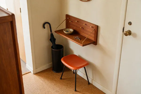 A small entryway with a wall-mounted walnut shelf, key hooks, a black umbrella stand, and a small orange stool with slender black legs. Cream walls and a cork floor create a warm, minimalist mid-century modern feel.