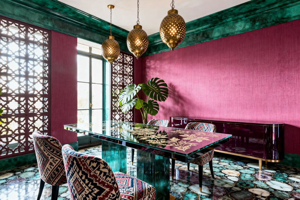 A jewel-toned dining room with fuchsia walls and deep green, marbleized moldings centers on a glass dining table featuring gold floral inlay and fluted emerald glass bases. Patterned upholstered chairs surround the table beneath three perforated brass pendant lights; a glossy plum credenza lines the wall, agate-slice stone flooring adds movement, geometric lattice panels flank black metal French doors, and a vase of monstera leaves sits on the table.