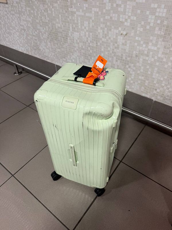 A heavily damaged silver Rimowa suitcase with multiple dents and scratches is shown on a luggage cart in an airport environment.