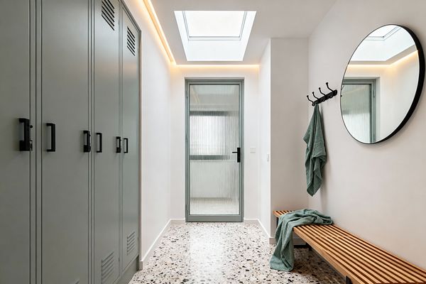 A bright contemporary mudroom features sage-gray built-in lockers with black handles on the left and a slatted oak bench with black legs on the right. Speckled terrazzo flooring leads to a ribbed-glass door under a skylight with warm cove lighting; a large round black-framed mirror and a row of black hooks hold muted green towels against white walls.