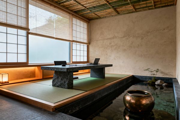 A tranquil Japanese Zen home office with a raised tatami platform sits beside a shallow reflecting pool edged in dark stone. A low granite desk with rough stone supports faces shoji windows filtered by woven bamboo blinds. Warm cove lighting washes light wood trim, while earthen plaster walls and a bamboo slat ceiling add soft texture. A legless floor chair, a small paper lantern, a bronze vessel, and a mossy rock vignette complete the minimal, nature-centered workspace.