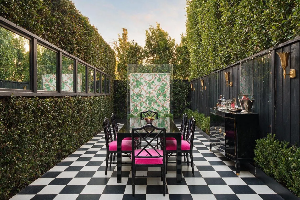 A narrow backyard dining area with a bold black-and-white checkerboard floor, surrounded by tall hedges and dark fencing. A black glass dining table with black chairs and bright pink cushions sits in the center, while a black sideboard and brass wall sconces line the right side, creating a polished Hollywood Regency look.