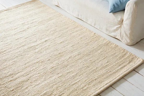 A light cream textured area rug lies on white painted floorboards beside a soft ivory sofa with a pale blue cushion. The rug has a woven, slightly ribbed natural fiber look that adds warmth and a relaxed coastal feel.