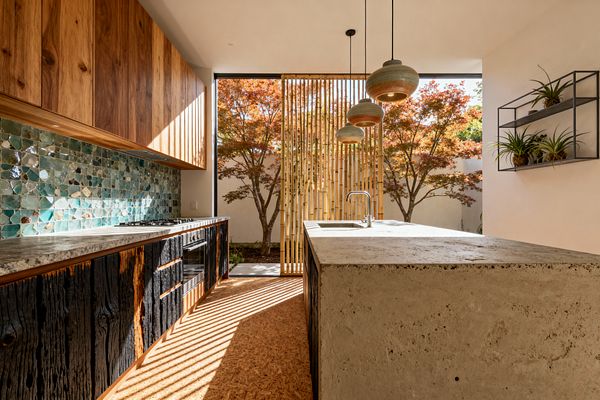 Sunlit kitchen with floor-to-ceiling opening to a courtyard of maple trees. A bamboo slat screen and three ceramic pendants hang over a cast-concrete island with an integrated sink; the run of cabinetry combines walnut uppers, shou sugi ban lower fronts, and a green zellige tile backsplash. Cork flooring and a black metal wall shelf with potted plants reinforce the biophilic palette of warm wood, stone, and living greenery.