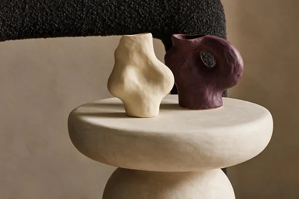 A modern decorative vignette shows two small sculptural ceramic vessels on a rounded pedestal table. One vessel is ivory and matte, while the other is deep plum with a cutout handle, set against a warm neutral background and a dark textured textile.