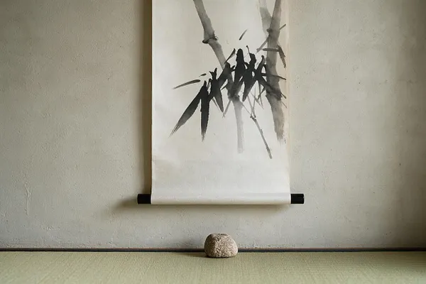 A minimalist Japanese Zen interior scene with a hanging ink scroll featuring expressive black bamboo brushwork on white paper. The scroll is mounted against a textured beige wall above a simple rounded stone placed on a pale tatami mat floor.