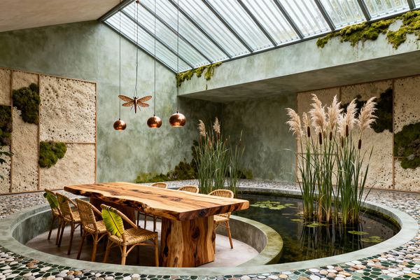 A biophilic dining room features a circular sunken seating area with a live-edge wooden table and rattan chairs with leaf‑green cushions, encircled by an indoor pond with lily pads, reeds, and tall grasses. Sage green limewash walls, porous stone panels inset with preserved moss, a pebble mosaic floor, and a large sloped glass skylight define the space; copper globe pendants and a dragonfly sculpture hang above, casting warm light.