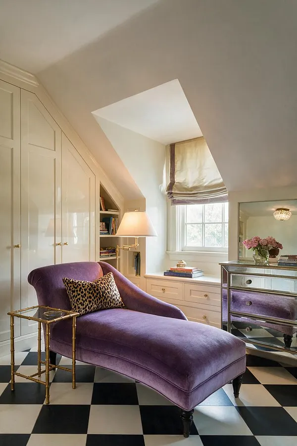 A Hollywood Regency master bedroom corner with a purple velvet chaise lounge, leopard-print accent pillow, and black-and-white checkered floor. Built-in cream wardrobes, a mirrored chest with a lamp, and a window seat with drawers create a chic, tailored look under a sloped ceiling.