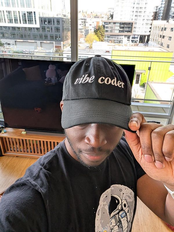 A person wearing a black cap with 'vibe coder' embroidered on it poses indoors.