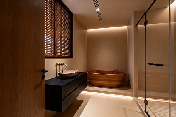 A contemporary bathroom with sand-toned plaster walls features a floating black vanity topped with a beige stone vessel and a wall-mounted brushed brass faucet beneath a perforated wood window screen. At the back, a freestanding wooden soaking tub sits on a slightly raised platform with concealed LED cove lighting, while a clear glass shower enclosure and minimal hardware complete the warm, streamlined space.