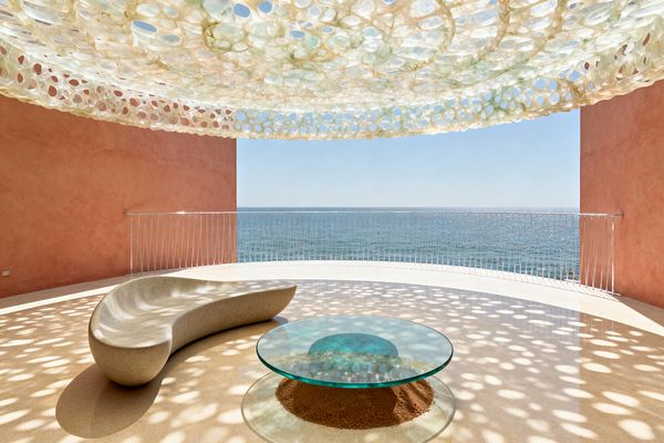 Open-air oceanfront patio with a curved white metal railing and expansive sea view. A perforated translucent canopy in milky white and pale green hangs overhead, casting circular shadows across a smooth sand-toned floor. At left, a sculptural pebble-gray concrete chaise has fluid contours, and at center a round clear-glass coffee table sits above a recessed ring of sand. Coral-colored stucco walls frame the bright, tranquil scene.