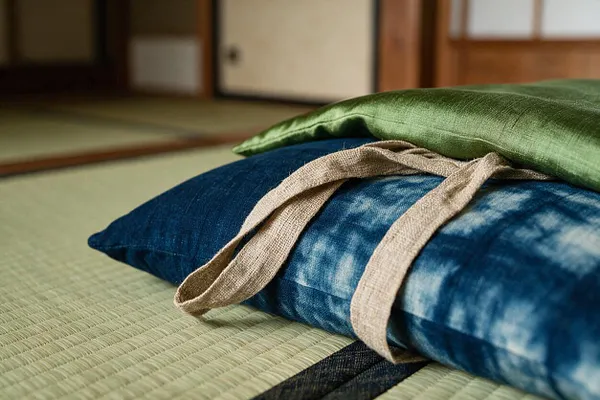 A low Japanese Zen interior with two stacked floor cushions resting on woven tatami mats. The top cushion is green silk-like fabric, the lower cushion is indigo-dyed textile tied with a natural beige linen strap, with shoji-style wood and paper doors softly blurred in the background.