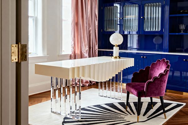 A Hollywood Regency home office features a cream lacquer desk with a scalloped edge and clear acrylic legs, topped by a white globe lamp with a marble column and brass base. Behind it, glossy cobalt blue built-ins with fluted glass doors and a white marble countertop pair with blush satin drapery, a plum velvet scallop-back chair with black legs and brass tips, a cream rug with a black Art Deco fan motif, warm herringbone wood flooring, and daylight from tall, white-trimmed windows.