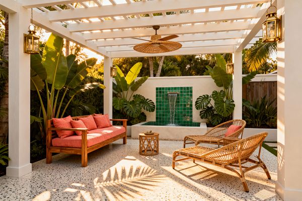Open-air tropical patio beneath a white pergola with a woven ceiling fan and brass lanterns. A long wood bench with coral cushions faces two rattan loungers and a round wicker side table on speckled terrazzo. At the back, a stucco wall features a waterfall over emerald square tiles into a small basin, surrounded by large banana leaves and monstera, all lit by warm late-afternoon sun.