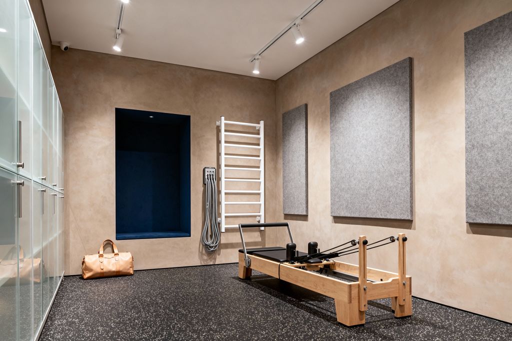A contemporary home gym with beige troweled plaster walls and black speckled rubber flooring is lit by ceiling track lights. A light oak Pilates reformer stands to the right beside three large gray acoustic panels; a white wall-mounted stall bar and coiled gray cables are nearby. On the left, frosted glass cabinets provide storage, and a deep navy recessed niche sits above a tan duffel bag.