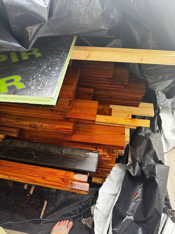 A stack of sauna wood is partially covered by a black tarp, showing signs of water exposure.