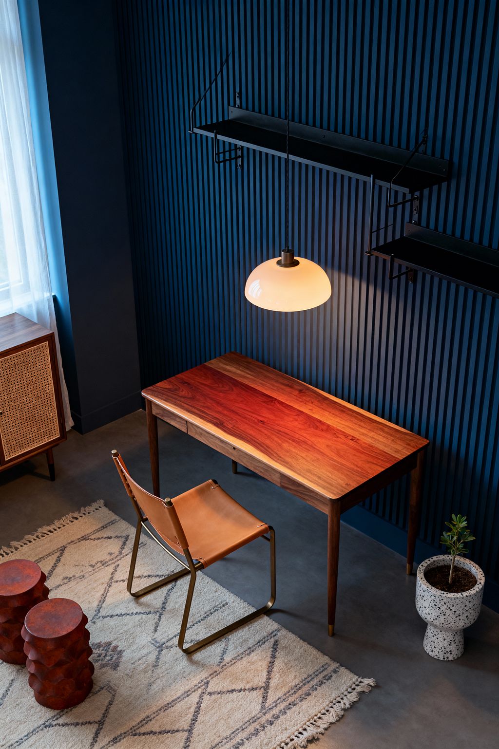 Mid-century modern home office featuring a slim walnut writing desk beneath a white glass dome pendant, set against deep navy vertical slatted paneling with black metal wall shelves. A tan leather sling chair sits on a cream rug with gray geometric lines over a gray concrete floor, with a terrazzo pedestal planter, two sculptural rust-toned stools, a rattan-front cabinet, and sheer curtains filtering light from a nearby window.