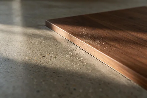 A close-up interior detail shows a thin walnut wood platform or slab hovering beside a polished concrete floor. Soft sunlight casts long shadows across the rough concrete, highlighting the contrast between the warm wood grain and the cool, textured surface.