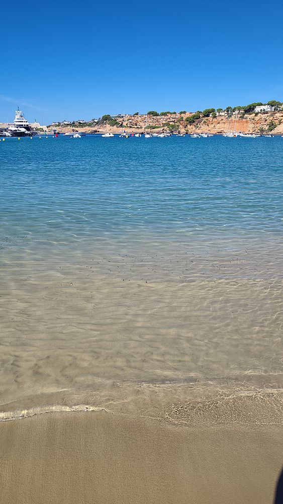 A clear and calm sandy beach with transparent water leading to a bay filled with boats and a coastal cliff with buildings under a clear blue sky.