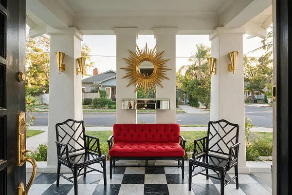 A front porch styled in Hollywood Regency fashion with a black-and-white checkerboard floor, a red tufted loveseat, and two black geometric chairs arranged symmetrically. White columns frame a gold sunburst mirror, mirrored console, and brass wall sconces, creating a dramatic, polished entrance.