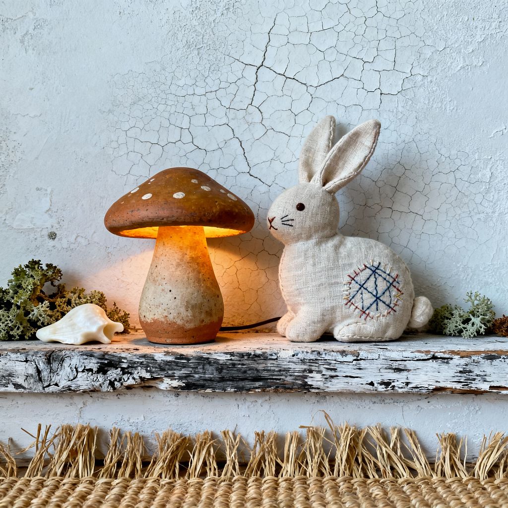 A close-up of a kids’ room vignette shows a rustic white-painted wood shelf with peeling finish set against a cracked plaster wall. A small ceramic mushroom lamp glows warmly beside a handmade linen rabbit plush with simple embroidery. Natural accents include clumps of lichen and a white seashell, while a woven jute rug with fringe appears along the bottom edge. The scene uses earthy textures and soft light to convey a calm, wabi-sabi mood.