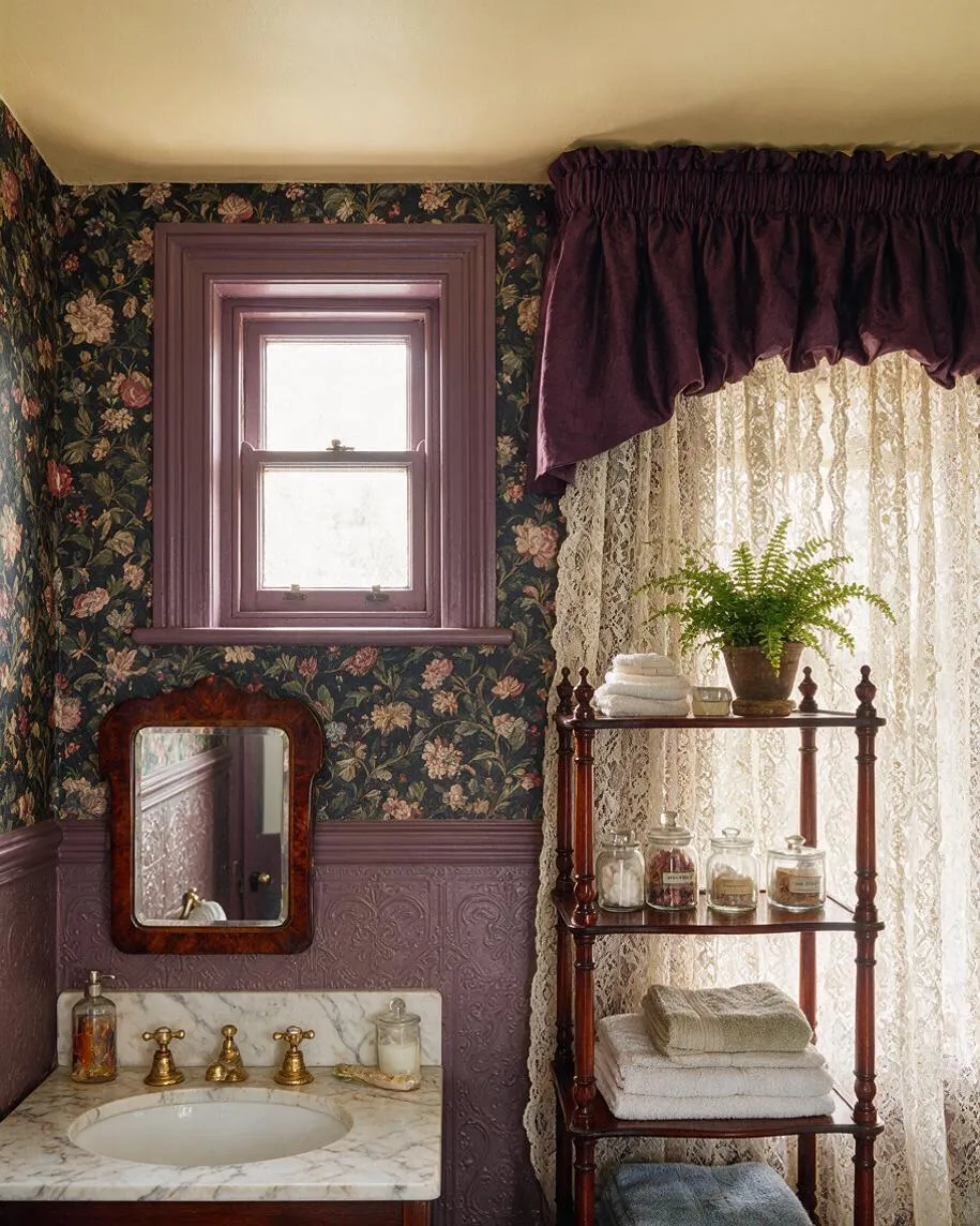 A Victorian-style master bathroom with dark floral wallpaper, mauve painted trim, and a marble sink vanity with brass faucets. Lace curtains and a plum draped valance soften the bright window, while a wooden étagère holds folded towels, glass jars, and a potted fern.