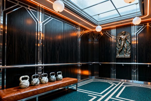An Art Deco home gym features dark wood wall panels with polished chrome geometric inlays and continuous metal handrails. A teal terrazzo floor with crisp white borders grounds a long cognac leather bench topped with marble kettlebells. A bronze bas-relief of an athlete decorates the wall, while opal globe pendants and a large frosted skylight illuminate the space; warm perimeter LEDs add an amber glow.