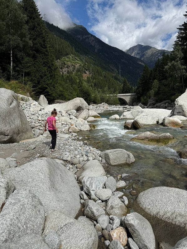 A person stands by a rocky riverbank surrounded by mountains and trees.
