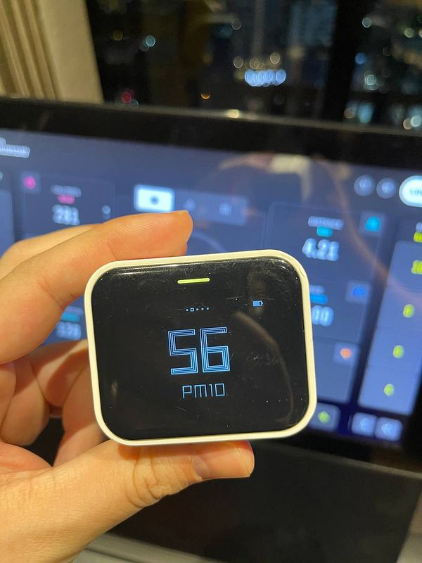 A hand holds a digital air quality monitor displaying a PM10 reading of 56.