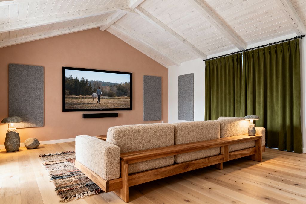 A Scandinavian media room features a terracotta plaster wall with a wall-mounted TV and slim black soundbar, flanked by gray felt acoustic panels. A solid oak sofa with beige bouclé cushions sits on light oak floors beside a striped flatweave rug; olive blackout curtains cover the window wall, a whitewashed vaulted wood ceiling spans overhead, and two stone lamps provide warm, soft accent lighting.