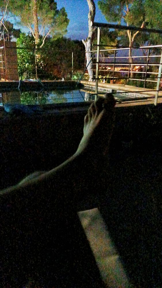 A pair of feet is resting near a poolside at night, surrounded by trees and a serene atmosphere.