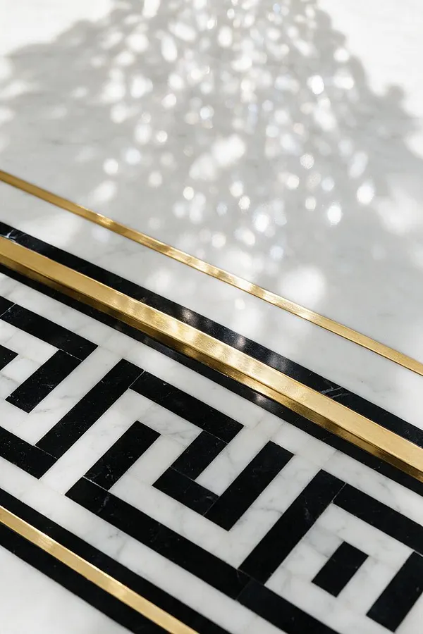 A close-up of a polished white marble surface with a bold black geometric inlay framed by slim brass strips. Soft reflected light creates a shimmering pattern across the stone, emphasizing the luxe contrast and decorative Hollywood Regency feel.