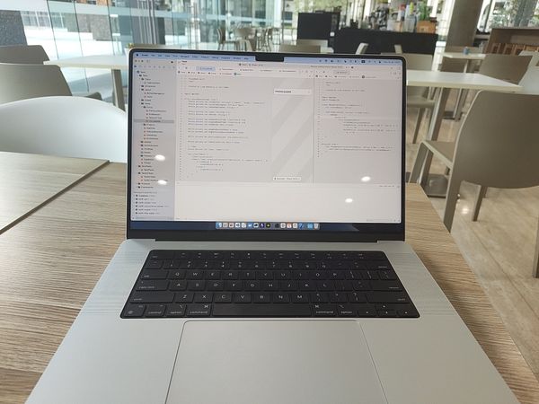 A laptop displaying a coding environment for Swift programming is set on a wooden table in a modern café.
