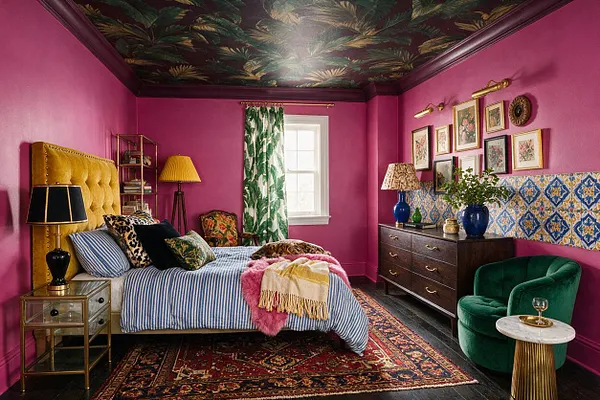 A colorful master bedroom with hot pink walls, a dark botanical ceiling, and a bed dressed in blue-and-white stripes with layered patterned pillows and throws. The room includes a mustard velvet headboard, mixed artwork on the walls, a dark wood dresser, a green velvet chair, and eclectic lamps and decor.