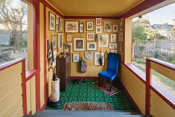 A small front porch styled in a maximalist way, with mustard yellow walls trimmed in deep red, a green patterned floor, and a dense gallery wall of framed artwork. A bright blue chair, umbrella stand, and layered doormats add color and personality, while the open sides reveal a fenced yard outside.