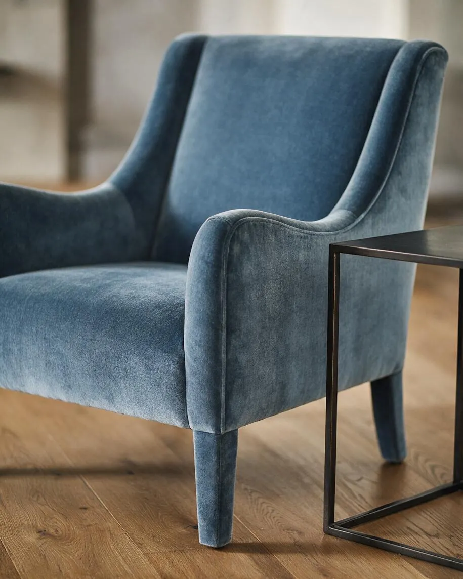 A transitional-style armchair in blue velvet with a gently curved back, padded arms, and tapered legs sits on a light wood floor. A slim black metal side table with a square frame is positioned beside it, creating a refined, modern-classic look.
