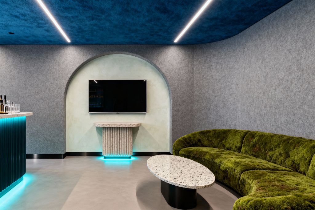 A contemporary media room features a curved moss-green velvet sectional facing a wall-mounted TV set within a pale plaster arched niche. An oval terrazzo coffee table and a fluted stone console with cyan underlighting sit on a light polished concrete floor, while gray felt acoustic wall panels and a deep cobalt-blue ceiling with linear LED strips create a softly lit atmosphere; a ribbed teal bar with a light stone top glows with matching LED at the left.