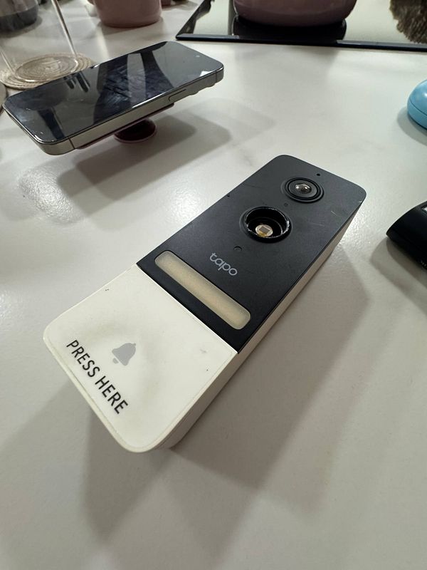 A Tapo doorbell device is shown on a table, with visible damage and a smartphone nearby.