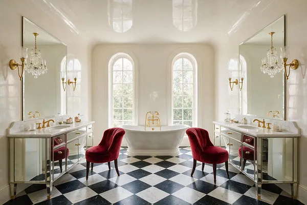 A luxurious master bathroom in Hollywood Regency style features twin mirrored vanities with marble countertops, gold fixtures, and crystal sconces on both sides. A sculptural white freestanding tub sits centered beneath two tall arched windows, with two deep red velvet accent chairs adding a dramatic touch over a black-and-white checkerboard floor.