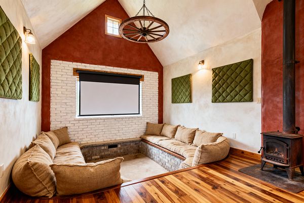 A farmhouse-style media room features a sunken U-shaped brick seating pit filled with oversized tan burlap cushions facing a roll-down projection screen set within a whitewashed brick wall. The vaulted room has terracotta red gable ends, warm plaster walls, olive green quilted acoustic panels, and a rustic wagon-wheel chandelier. A cast-iron wood stove rests on a hearth beside striped hardwood flooring, with glass-shaded wall sconces providing soft light.