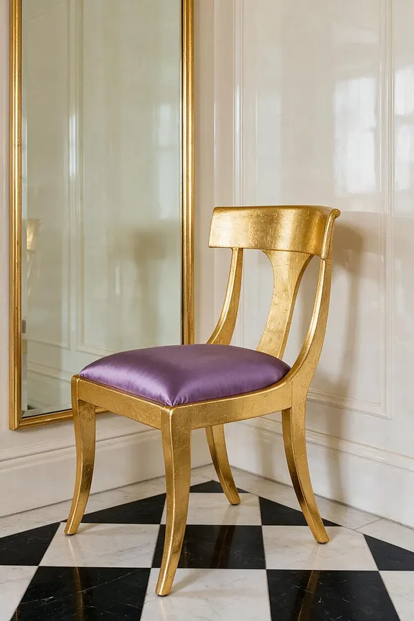 A single Hollywood Regency chair stands in a refined room with white paneled walls and a black-and-white checkered marble floor. The chair has a gold leaf frame, a curved open back, and a plush purple satin seat cushion, with a tall mirror behind it reflecting light.
