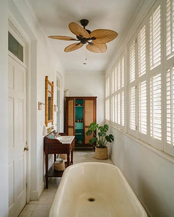 A long tropical-style master bathroom with white walls, a cream freestanding bathtub, and a dark wood vanity along one side. Large shuttered windows line the opposite wall, while a bamboo-framed mirror, woven cabinet doors, rattan accents, and a leafy potted plant add warm natural texture.