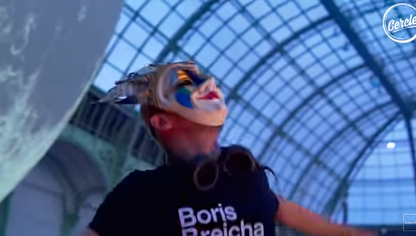 A performer wearing a colorful mask and a t-shirt with 'Boris Brejcha' printed on it is dancing energetically under a glass dome structure.