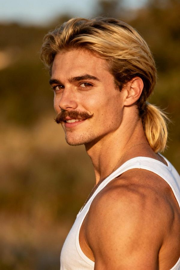 A young man with long blonde hair and a mustache poses confidently outdoors.