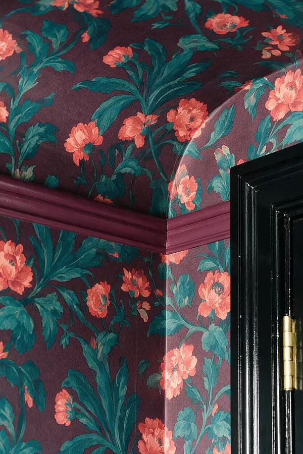 A close-up of a room corner covered in bold floral wallpaper with coral blossoms and teal leaves on a deep plum background. A glossy burgundy chair rail wraps the corner, and part of a black door with a brass hinge appears at the right edge.