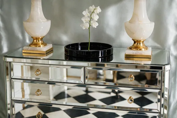 A mirrored console table with a glass top sits against a soft silver backdrop, styled with two ivory-and-gold urn lamps and a black tray holding a white orchid. The reflective drawer fronts, brass ring pulls, and black-and-white checkered floor create a polished Hollywood Regency look.