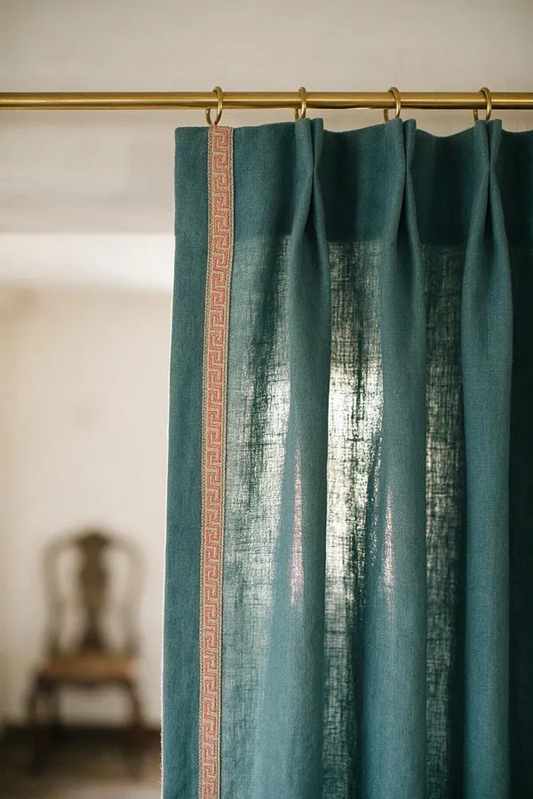 A close-up of teal curtains hanging from a brass rod with round rings. A narrow pink-and-cream Greek key trim runs vertically near the edge, while a blurred chair and pale wall appear in the background.